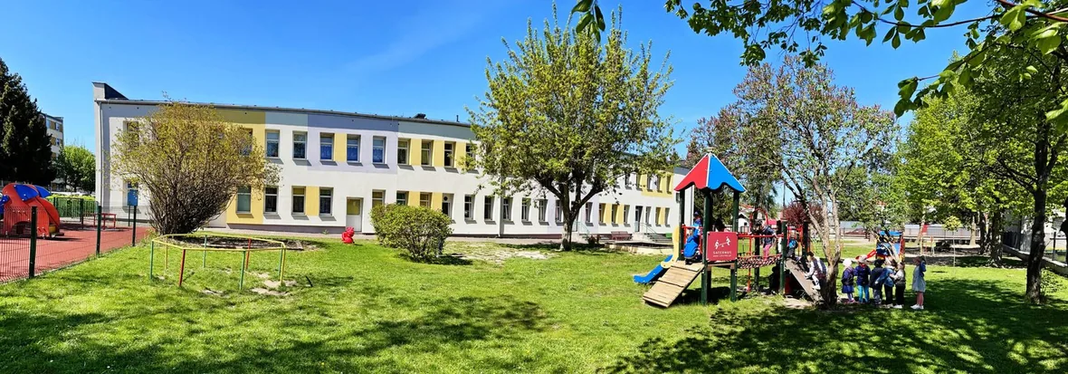 Budynek przedszkola od strony ogrodu: parterowo-piętrowy obiekt z jasną elewacją, przed nim trawnik i plac zabaw z kolorowymi urządzeniami.