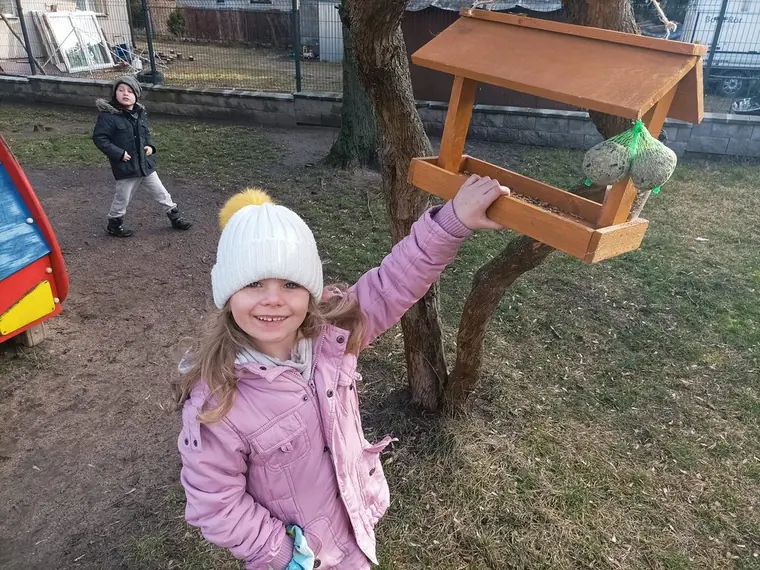 Dziewczynka w różowej kurtce i białej czapce zawiesza kulę tłuszczową przy drewnianym karmniku przymocowanym do drzewa.