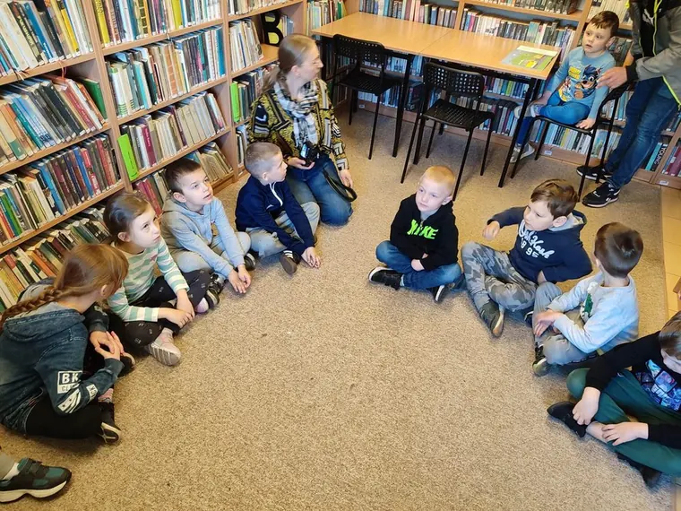 Dzieci siedzą w kręgu na podłodze między regałami z książkami podczas wizyty w bibliotece.