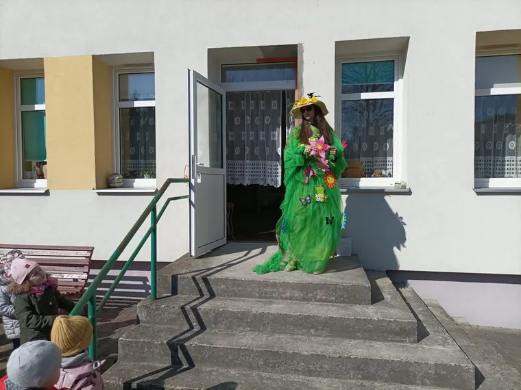 Osoba w zielonym stroju Pani Wiosny, ozdobionym kwiatami, stoi w drzwiach przedszkola; dzieci czekają na schodach.