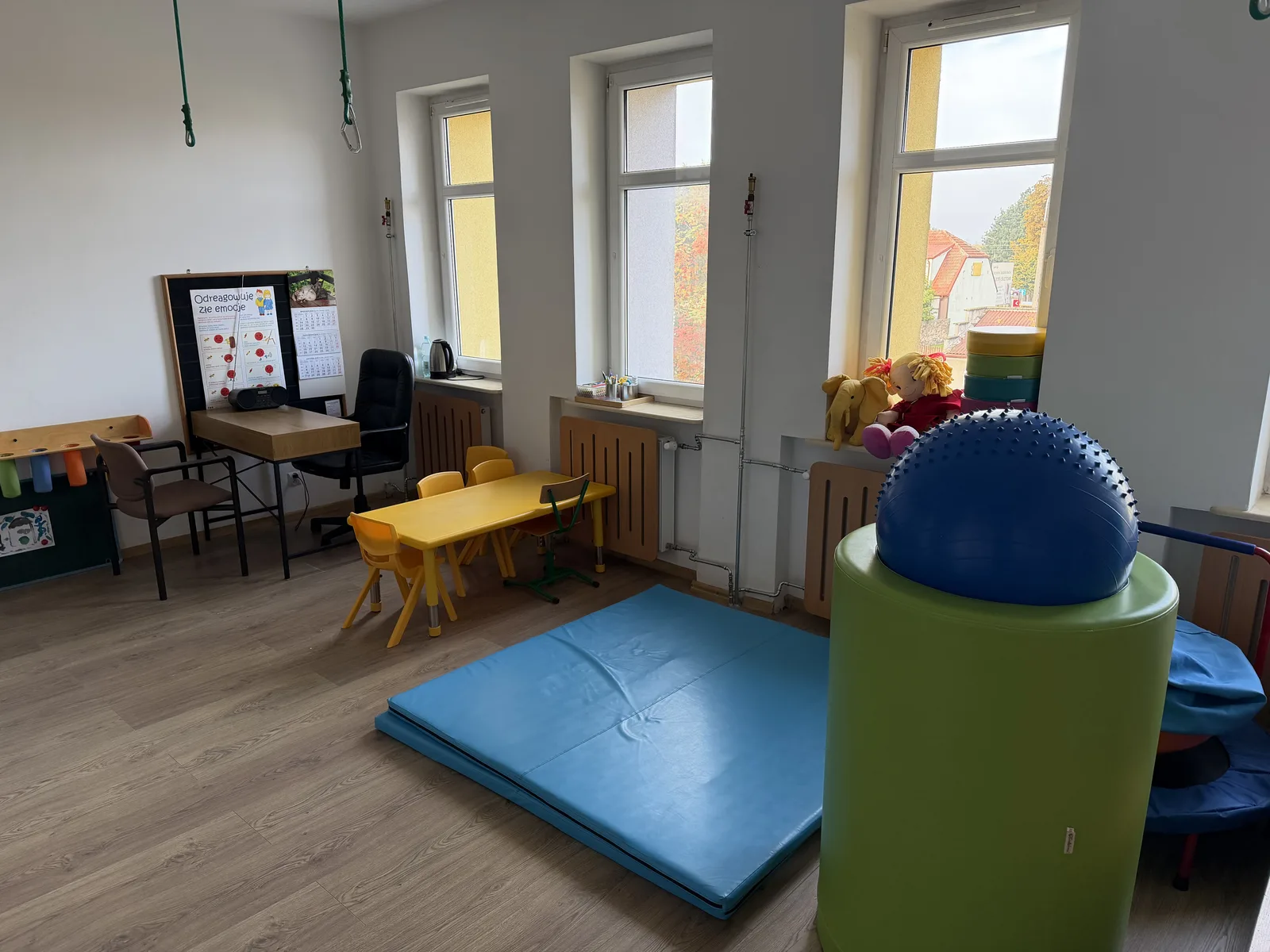Gabinet Integracji Sensorycznej: materac do ćwiczeń, duża piłka rehabilitacyjna na wałku, podwieszki u sufitu, stolik z krzesełkami i tablica z planszą o emocjach.