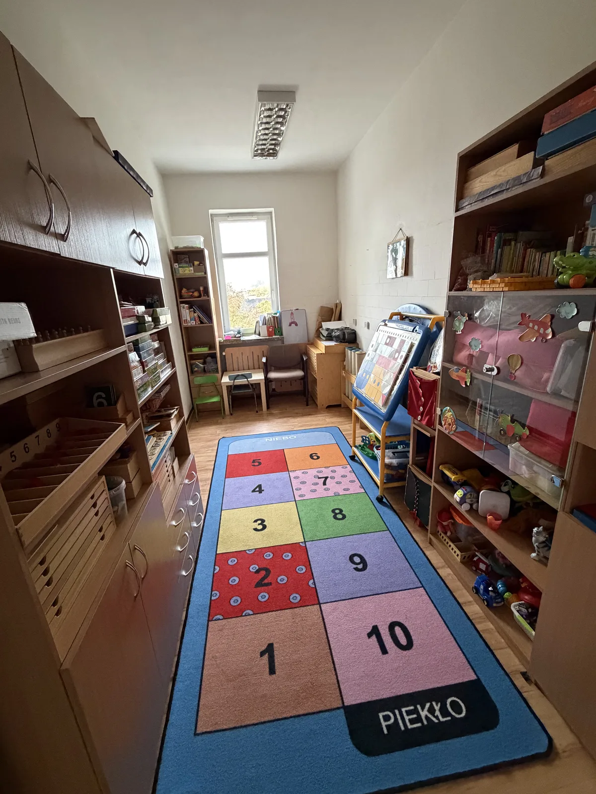Sala Montessori: dywan w formie gry w klasy z cyframi od 1 do 10, obok regały z pomocami do nauki liczenia i zabawkami.